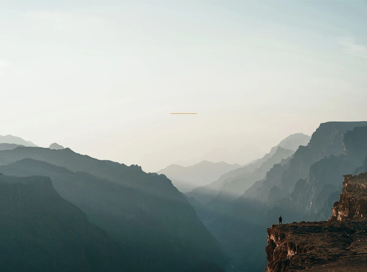 A lone figure on a cliff edge above layered mountain ridges at dusk